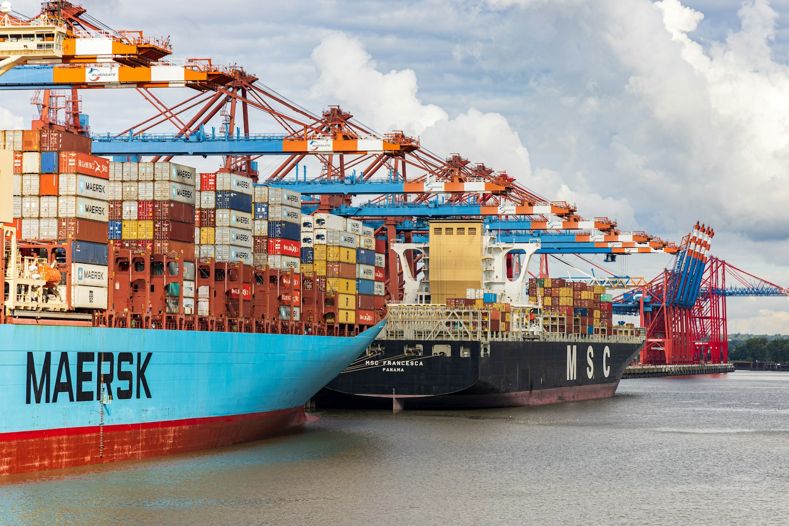 A laden container ship berthed under gantry cranes.